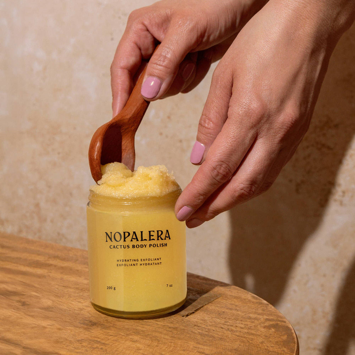 Person using Nopalera cactus body polish with a wooden spoon on a wooden surface.