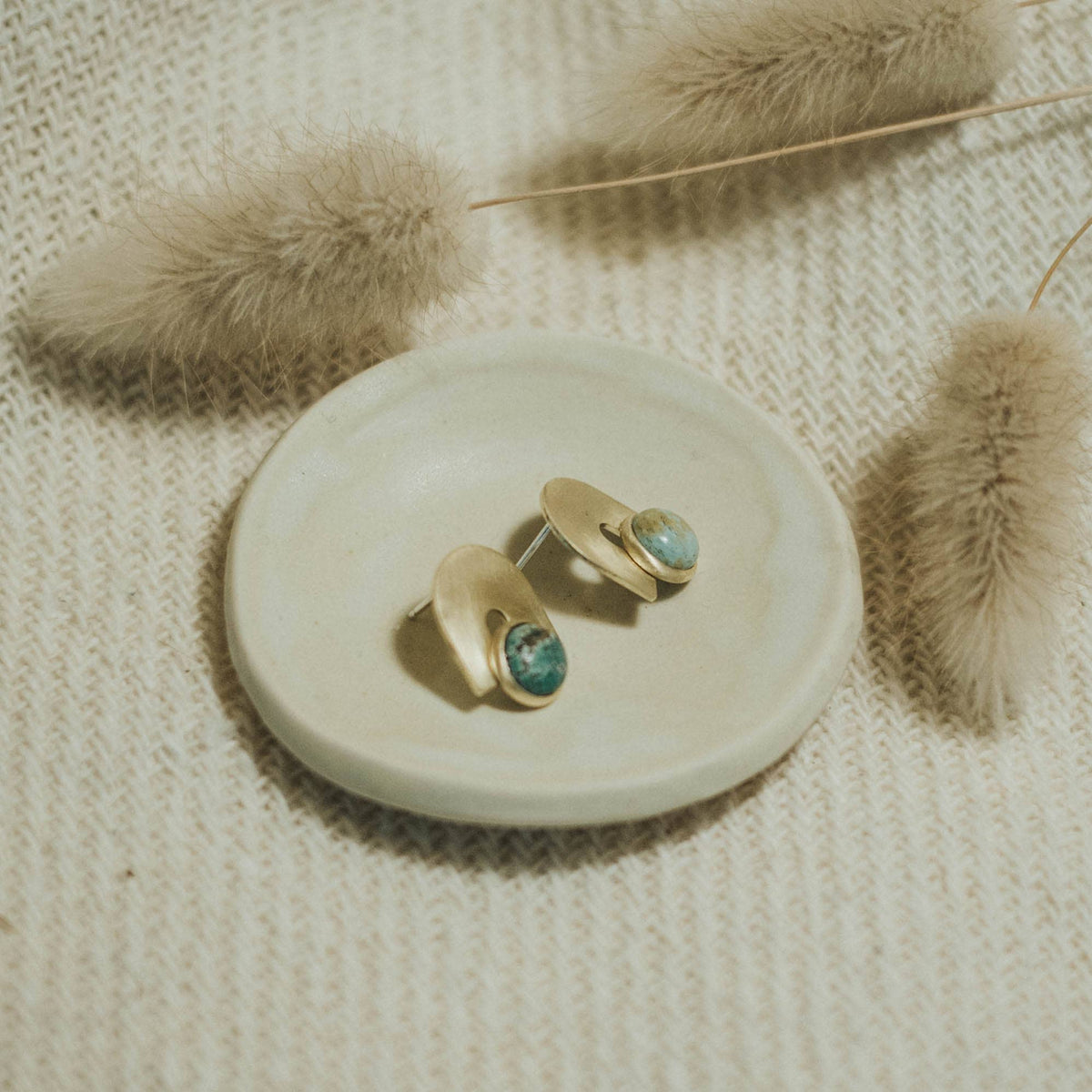 Gold earrings with turquoise stones on a white dish, surrounded by dried plants on a textured beige surface.