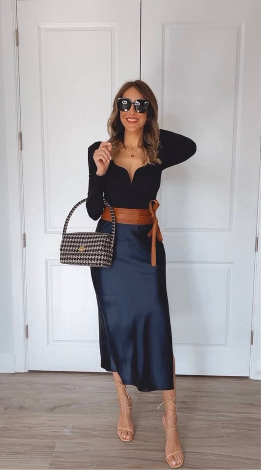 Woman in a black top and navy skirt with tan wrap belt, sunglasses and a handbag indoors.