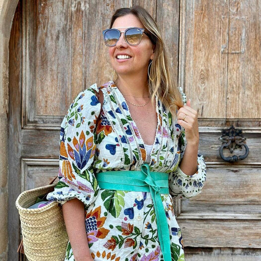 Woman wearing a colorful floral dress with an aqua belt, standing in front of a wooden door.