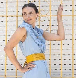 Woman in a light blue dress with a yellow belt standing against a white wall with yellow grid patterns.