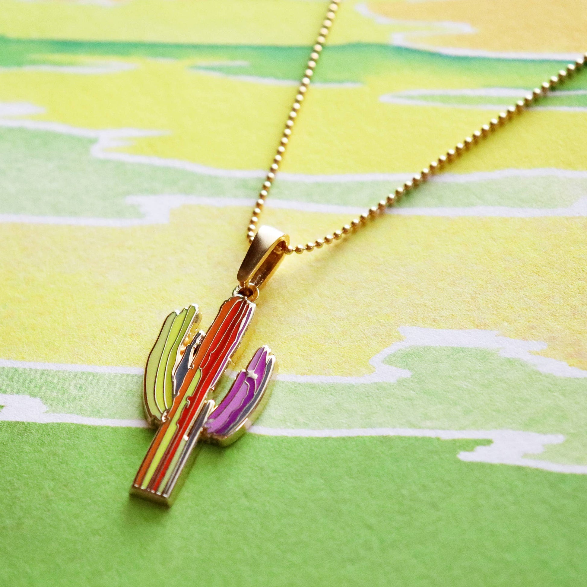 Colorful cactus-shaped pendant on a gold chain against a green and yellow patterned background